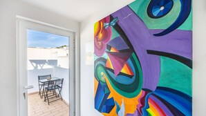 A room with a colorful abstract painting on the wall, a door leading to an outdoor area with a table and chairs, and a view of buildings in the distance. - Casa Da Paz and Love (Salema)