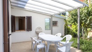 Eine überdachte Terrasse mit einem weißen Tisch und Stühlen, ein Fenster mit Rollläden und eine an der Wand befestigte Uhr.