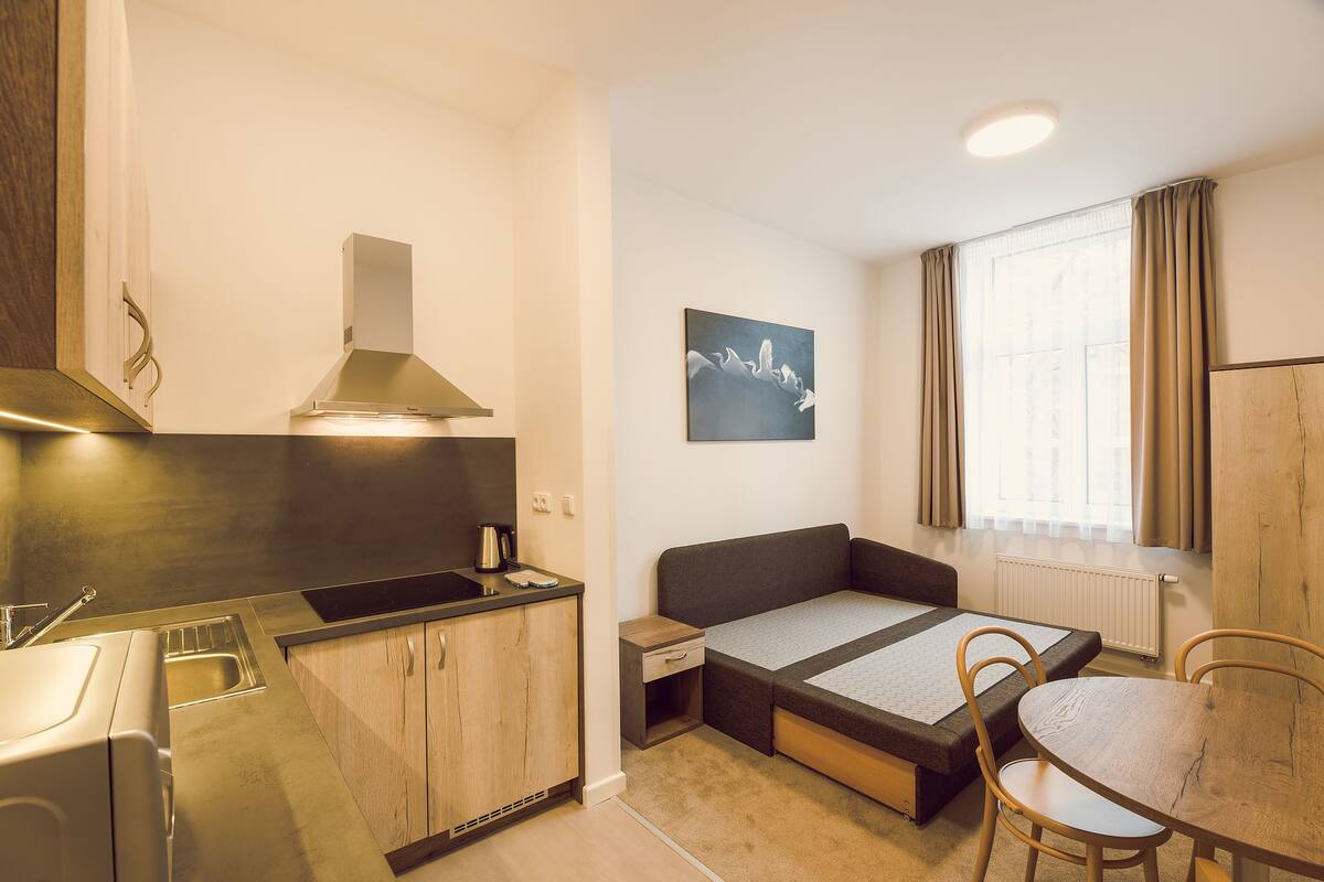 Habitación compacta con kitchenette, cama con mesa plegable y ventana con cortinas.