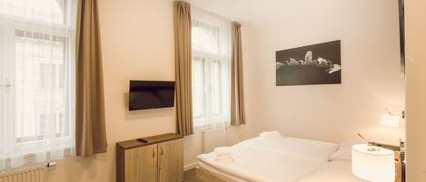 Habitación de hotel con una cama, televisión montada en la pared, armario y ventana con cortinas.