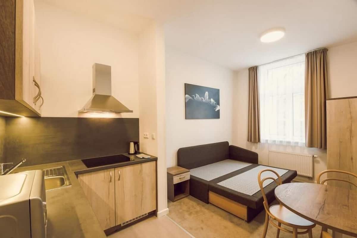 Una habitación compacta con kitchenette, una cama con espacio de almacenamiento, una mesa de comedor y un ventanal con cortinas.