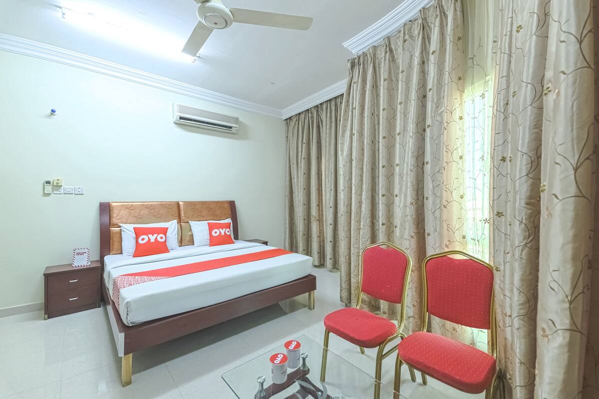 A hotel room with a bed, two chairs, a nightstand, an air conditioner, and a ceiling fan.