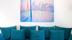 A teal-colored sofa with multiple cushions against a white wall, with a colorful painting above it.