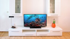 A modern living room with a flat-screen TV on a white entertainment unit, a small table with decorative items, and a glass cabinet with shelves.