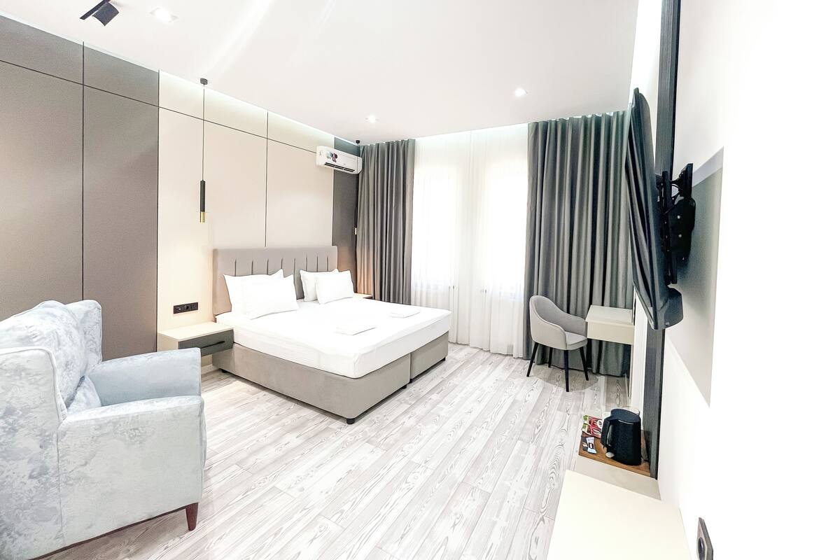 A modern hotel room with a large bed, a sofa, a desk, and a flat-screen TV mounted on the wall.
