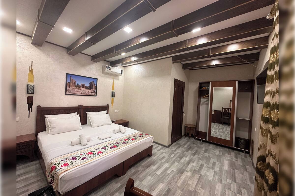 A hotel room with a large bed, wooden headboard, and a decorative bedspread. There is a mirror on the wall and a door leading to a bathroom.