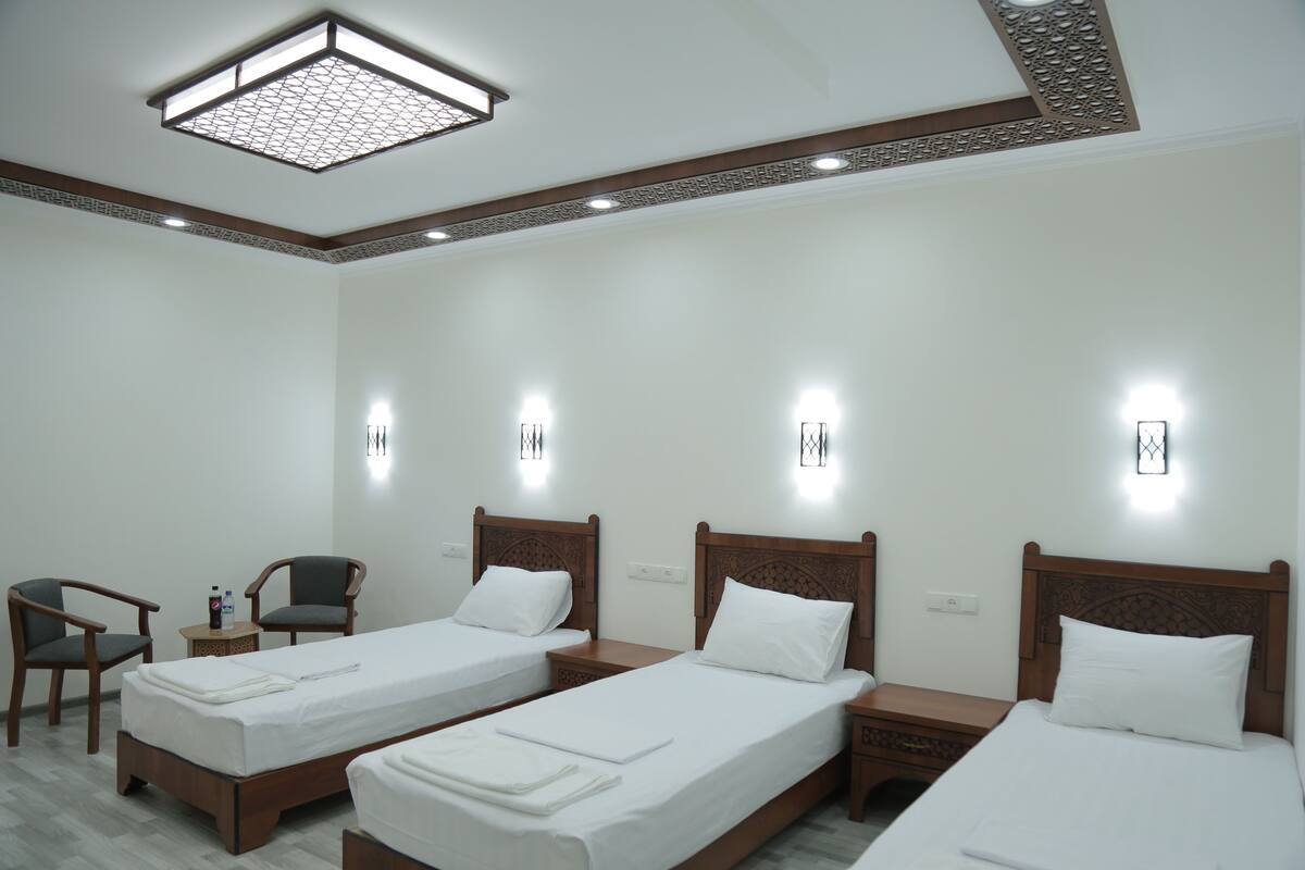 A hotel room with three single beds, wooden headboards, a small table with a chair, and wall-mounted lights.