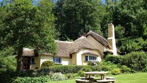 Een traditioneel huis met rieten dak en schoorsteen, omringd door weelderig groen en een picknicktafel op de voorgrond.