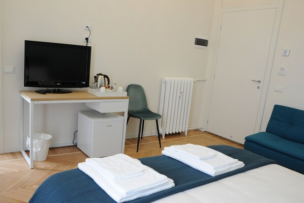 A hotel room with a bed, a desk with a computer, a chair, a radiator, and a blue sofa.