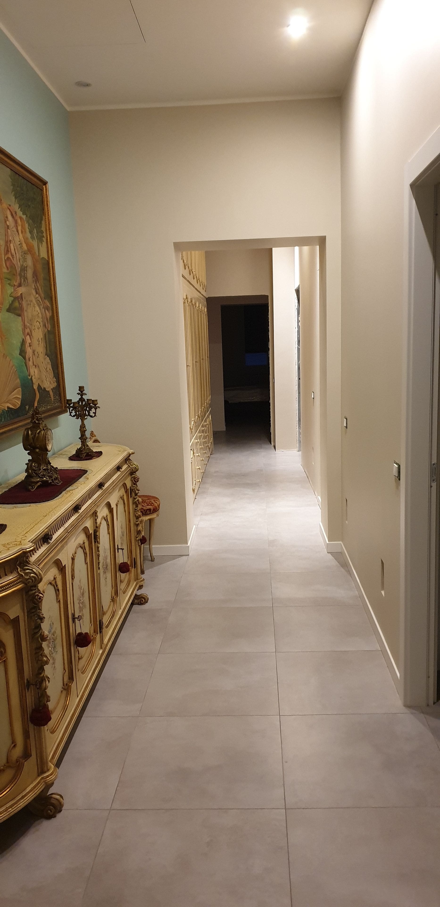 A hallway with a cream-colored floor, a decorative gold cabinet with red accents, and a painting on the wall.