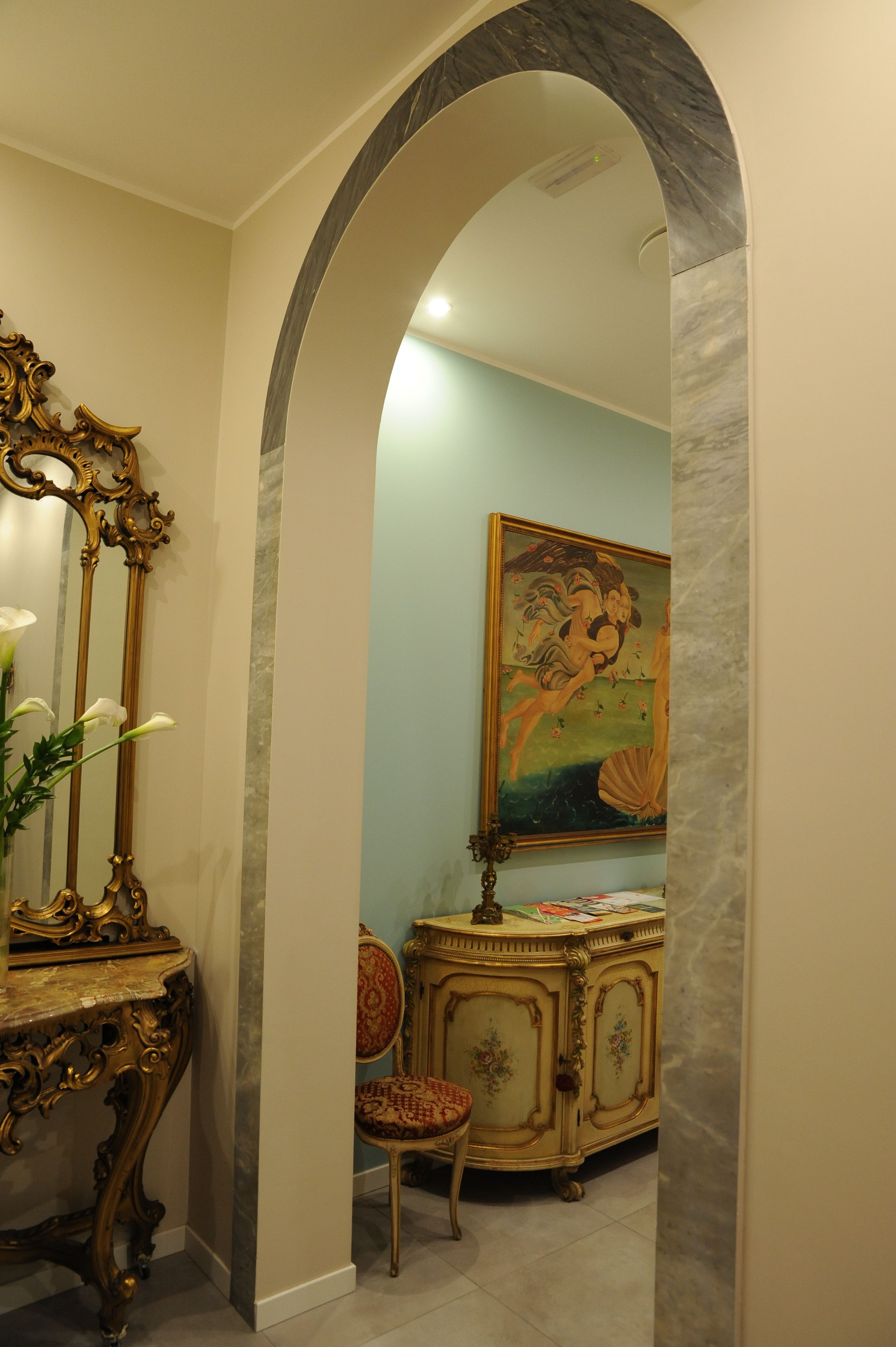 Un elegante corridoio con un arco in marmo, uno specchio con cornice dorata, una poltrona decorata con motivi floreali e un mobile con un quadro sopra.