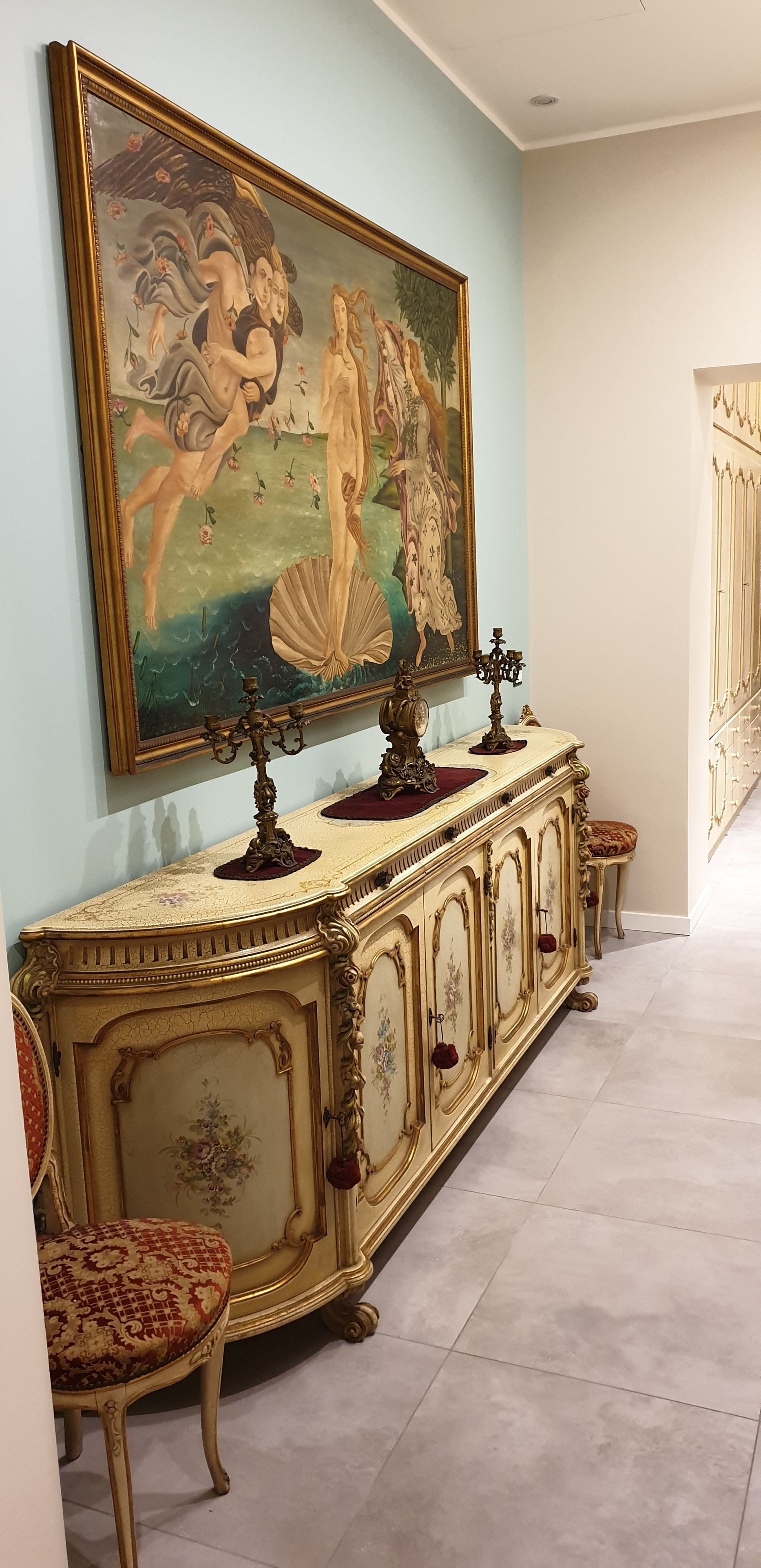 A grand hallway with a large classical painting, ornate furniture, and a decorative candelabra.
