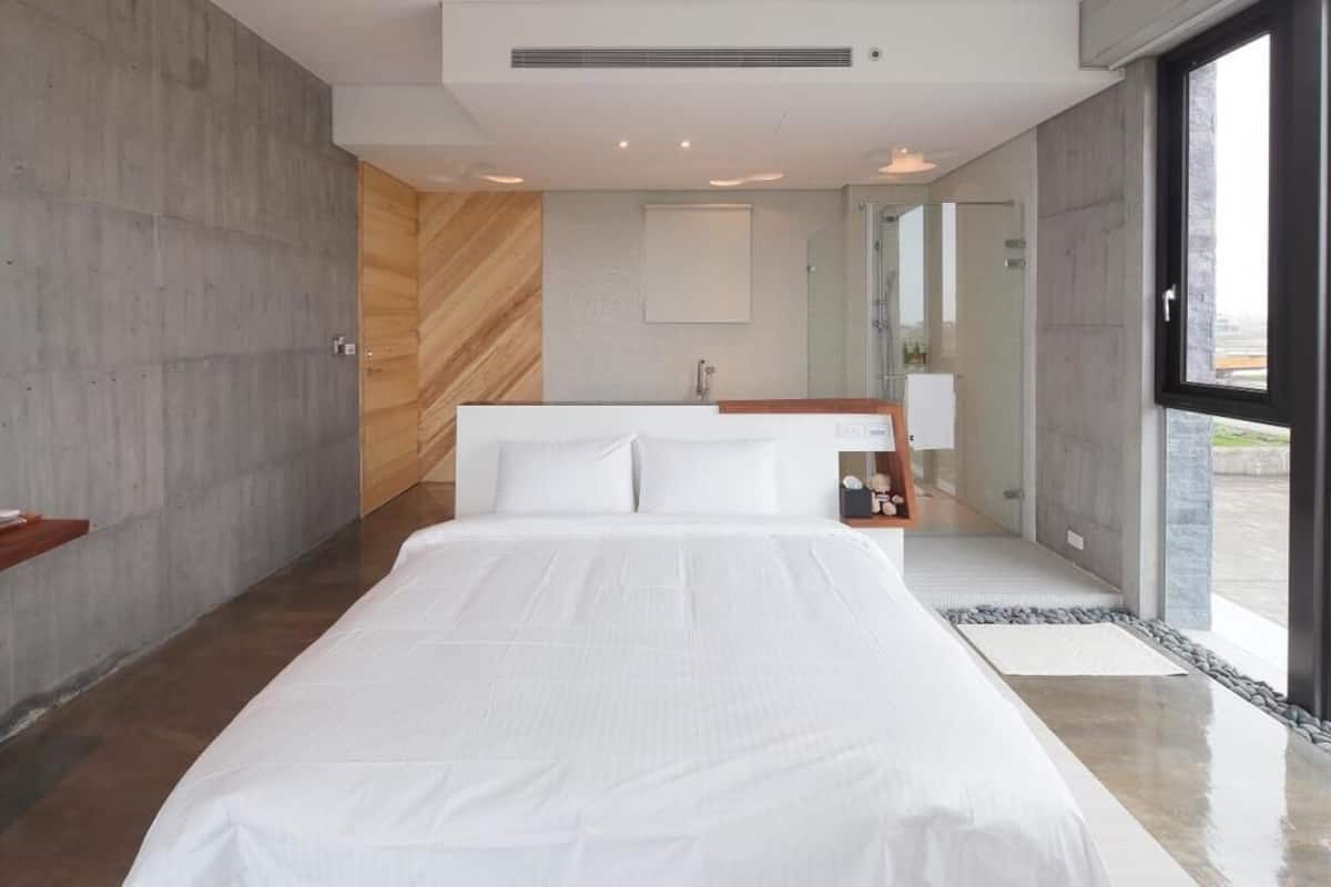 A modern bedroom with a large bed, a wooden accent wall, a built-in shower, and a view through a large window.