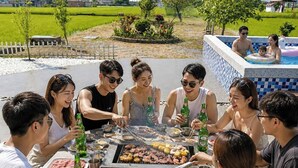 Eine Gruppe von Menschen versammelt sich um einen Grill mit verschiedenen Fleischsorten und Gemüsesorten, im Hintergrund ist ein Pool und eine gebirgige Landschaft zu sehen.