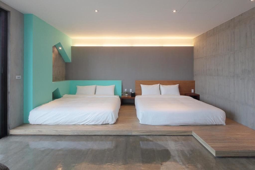 A modern hotel room with two beds, a wooden platform, and a concrete wall.