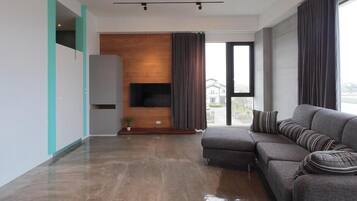 A modern living room with a grey sectional sofa, a flat-screen TV mounted on a wooden wall, and a large window with curtains.