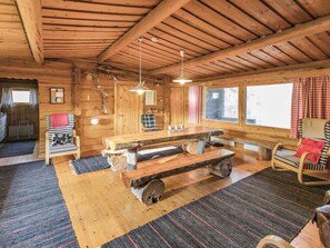 A rustic cabin interior with a wooden dining table, benches, a rocking chair, and a fireplace.