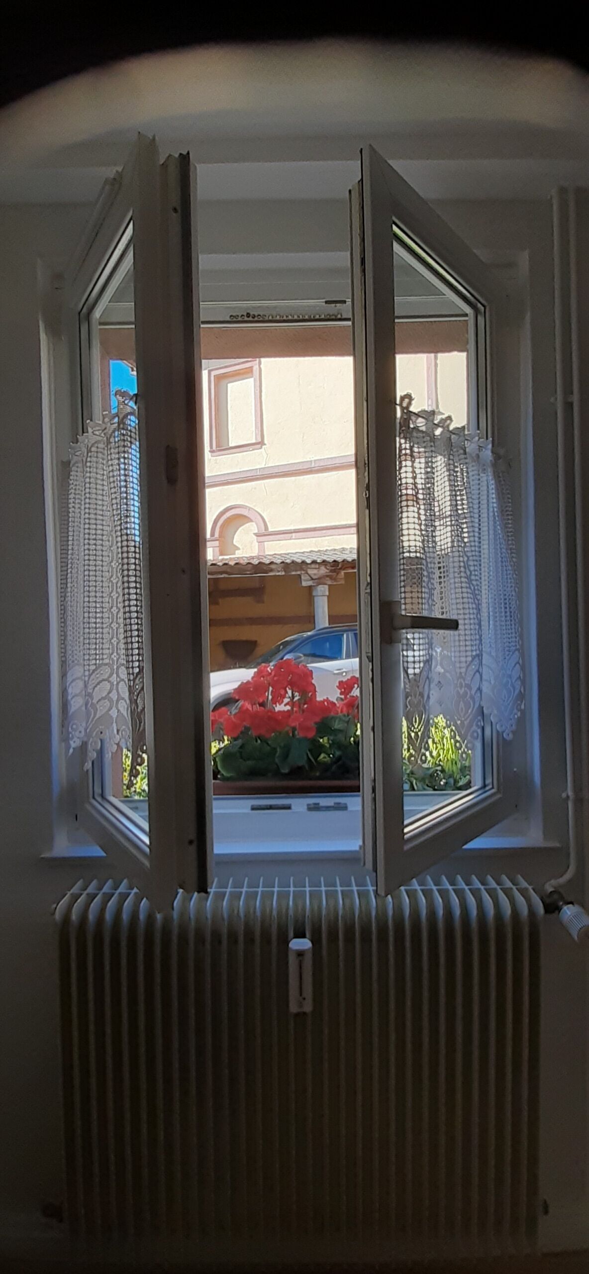 Ventana abierta con vista al exterior de un edificio, un radiador debajo y una cortina a la izquierda.