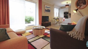 A living room with a sofa, a coffee table, and a TV. There is a dining area with chairs and a table. The room has a large window and a door leading outside.