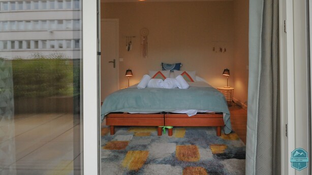 A bedroom with a bed, bedside lamps, a patterned rug, and a view of a building outside.