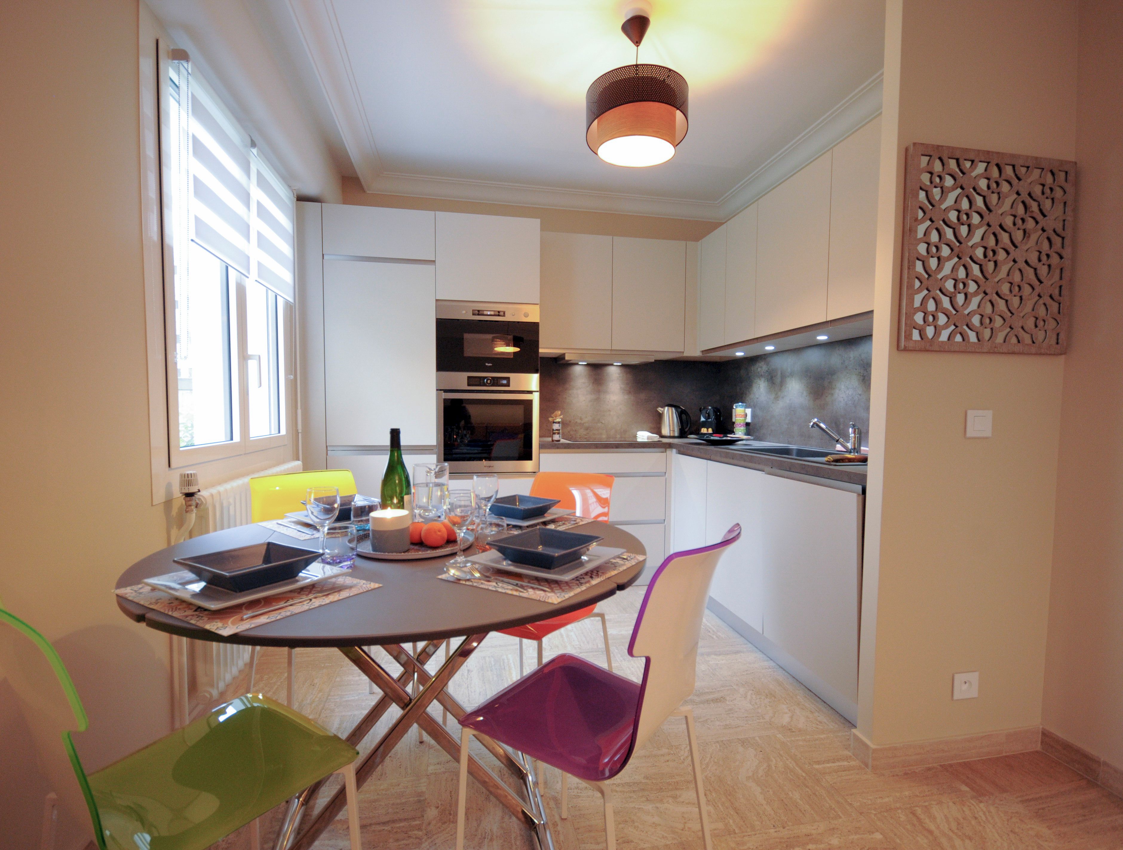 A modern kitchen with white cabinets, a built-in oven, and a dining area with a round table and colorful chairs.