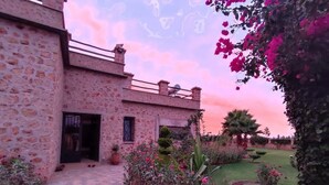 A stone building with a paved entrance and a garden with pink flowers and a palm tree. - Luxury villa with private pool not overlooked. (Fès)