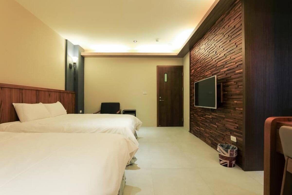 Habitación de hotel con dos camas, un televisor montado en una pared de ladrillo, una puerta y un pequeño cubo de basura.