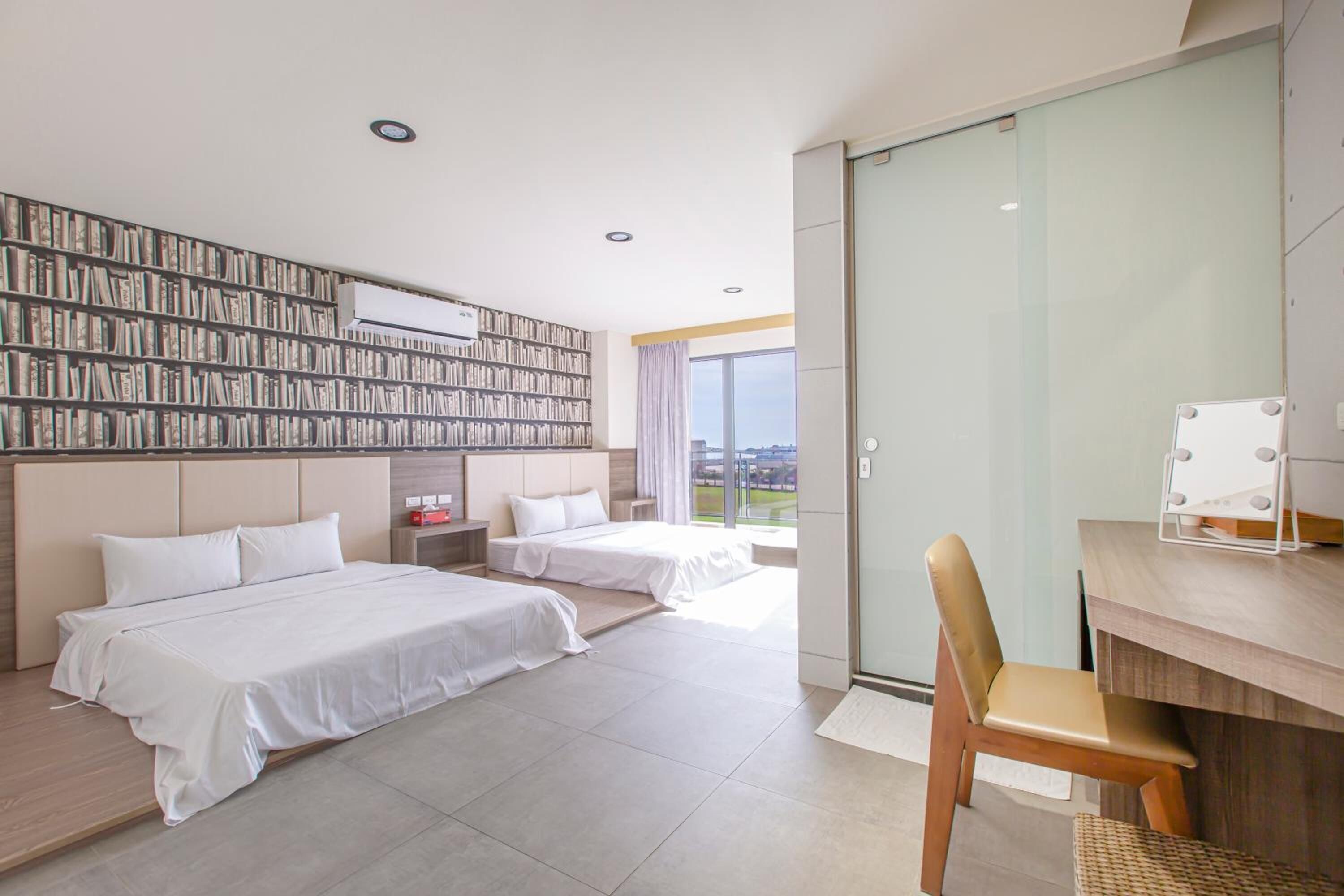 A modern hotel room with two beds, a bookshelf, a desk with a chair, and a large window offering a city view.