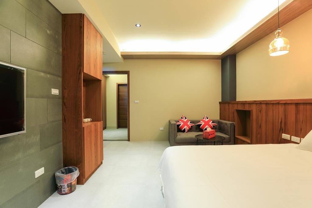 A modern hotel room with a large bed, a flat-screen TV mounted on the wall, a wooden wardrobe, a sofa, and a small table with red pillows.