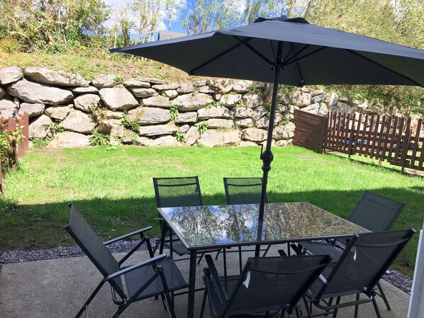 Een terras met een glazen tafel en stoelen, een parasol, een stenen muur en een houten hek.