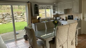 A kitchen with a dining area, featuring a glass table, chairs, and a view of a garden through a sliding door. - Cosy 3 bed holiday home (Kenmare)