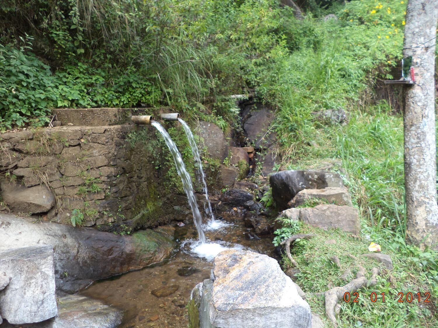 A small natural water feature with a stone wall and a metal pipe, surrounded by rocks and greenery.