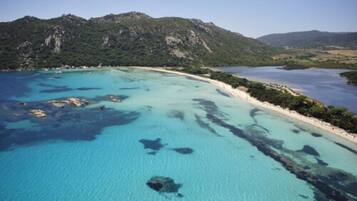 An aerial view of a coastal area with clear turquoise waters, a sandy beach, and a mountainous backdrop.