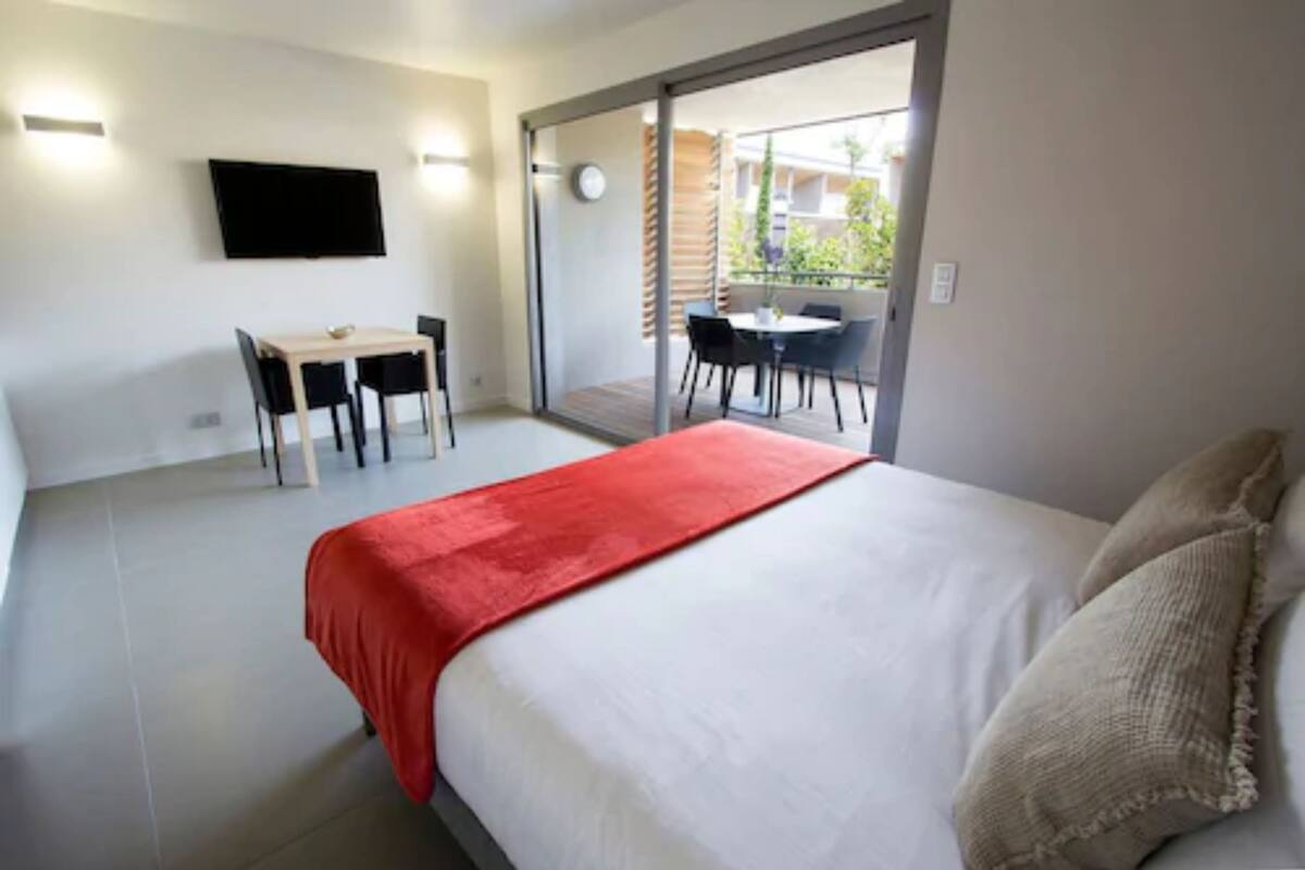 A bedroom with a bed, a desk, and a chair. There is a sliding glass door leading to an outdoor area with a table and chairs.
