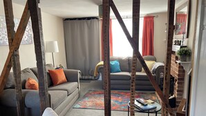 A living room with a grey sofa, a white armchair, and a coffee table with books. There is a brick fireplace and a window with curtains.