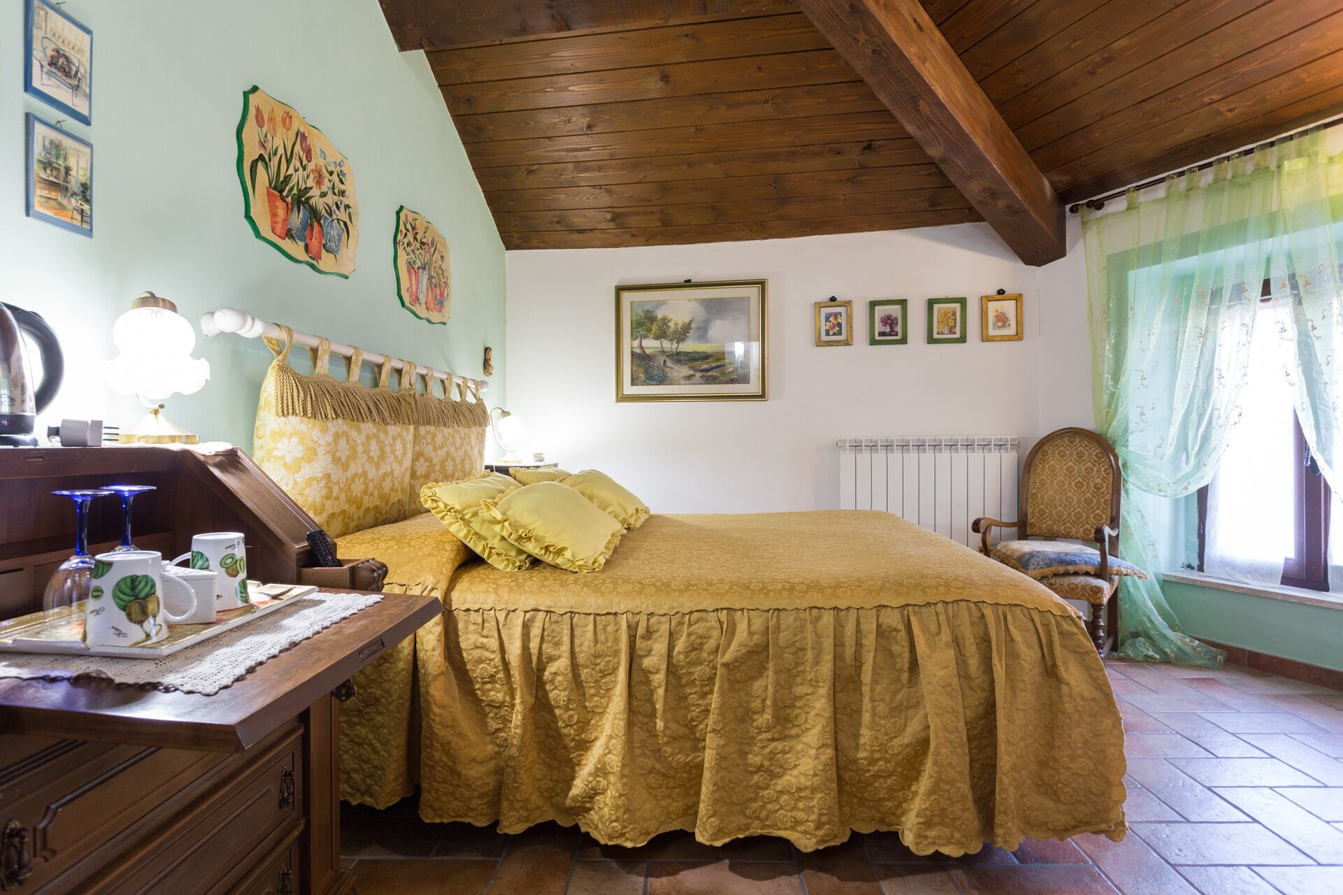 Une chambre confortable avec un plafond en bois, un lit recouvert d’une couverture jaune, une table de chevet en bois, une chaise et une fenêtre ornée d’un rideau vert.