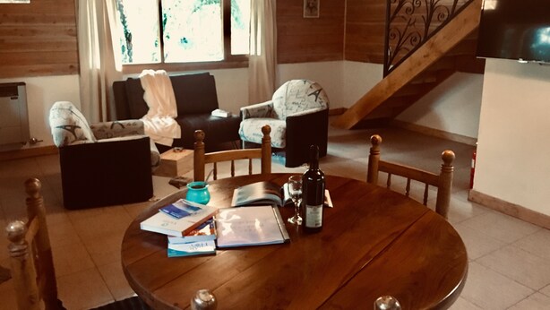 Un accogliente soggiorno con un tavolo da pranzo in legno, due divani, un tavolino da caffè con libri e una bottiglia di vino, e una scala che conduce al piano superiore.