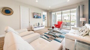 A modern living room with white sofas, a glass coffee table, and a red chair. There is a TV mounted on the wall and a sliding glass door leading to an outdoor area.