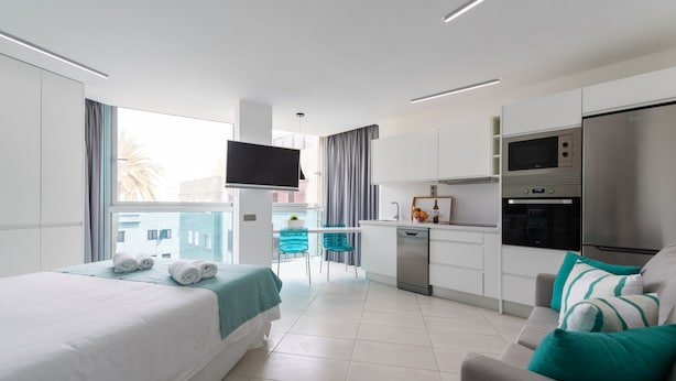 Una habitación de hotel moderna con una cama, un sofá, un televisor, una cocina con microondas y refrigerador, y un balcón con vistas a edificios.