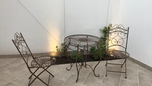 Un espace patio avec une table en métal et deux chaises, entouré de plantes en pot et d’un mur blanc.