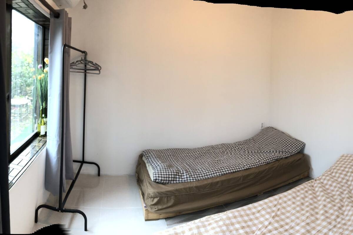 Una habitación pequeña y funcional con cama, ventana con cortina, espejo y estante con fotografías.