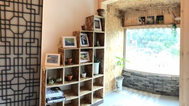 A room with a wooden bookshelf filled with framed photos and potted plants, a large window with a view of greenery, and a stone wall.