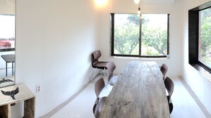 A dining area with a wooden table and chairs, a large window with a view of greenery, and a wall-mounted mirror.
