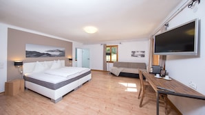 A modern hotel room with a large bed, a sofa, a desk with a flat-screen TV, and a wooden floor. - Alpenhaus Kesselfall (Kaprun)