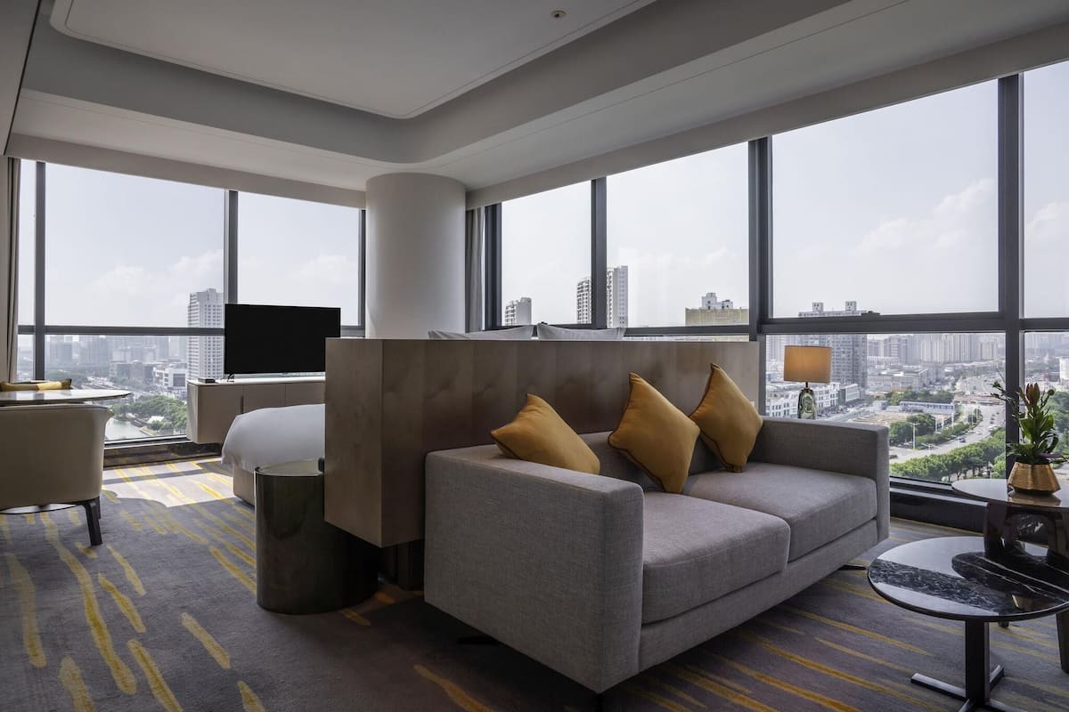 Una habitación de hotel moderna con un ventanal grande, un sofá, un televisor y una mesita.