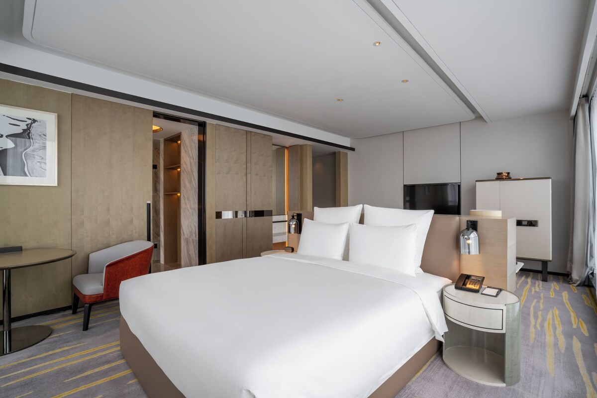 A hotel room with a large bed, a desk, a chair, a wardrobe, and a nightstand with a lamp.