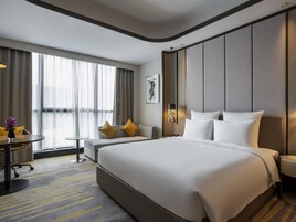 A modern hotel room with a large bed, a desk, and a chair. There is a window with curtains, a painting on the wall, and a lamp.