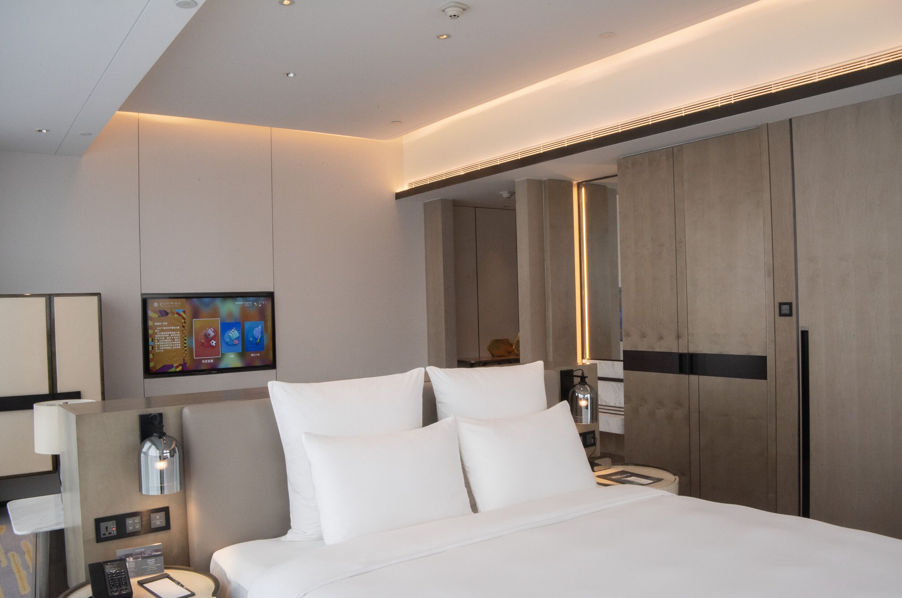 A modern hotel room with a large bed, a bedside table, a lamp, a wall-mounted TV, and a wooden wardrobe.