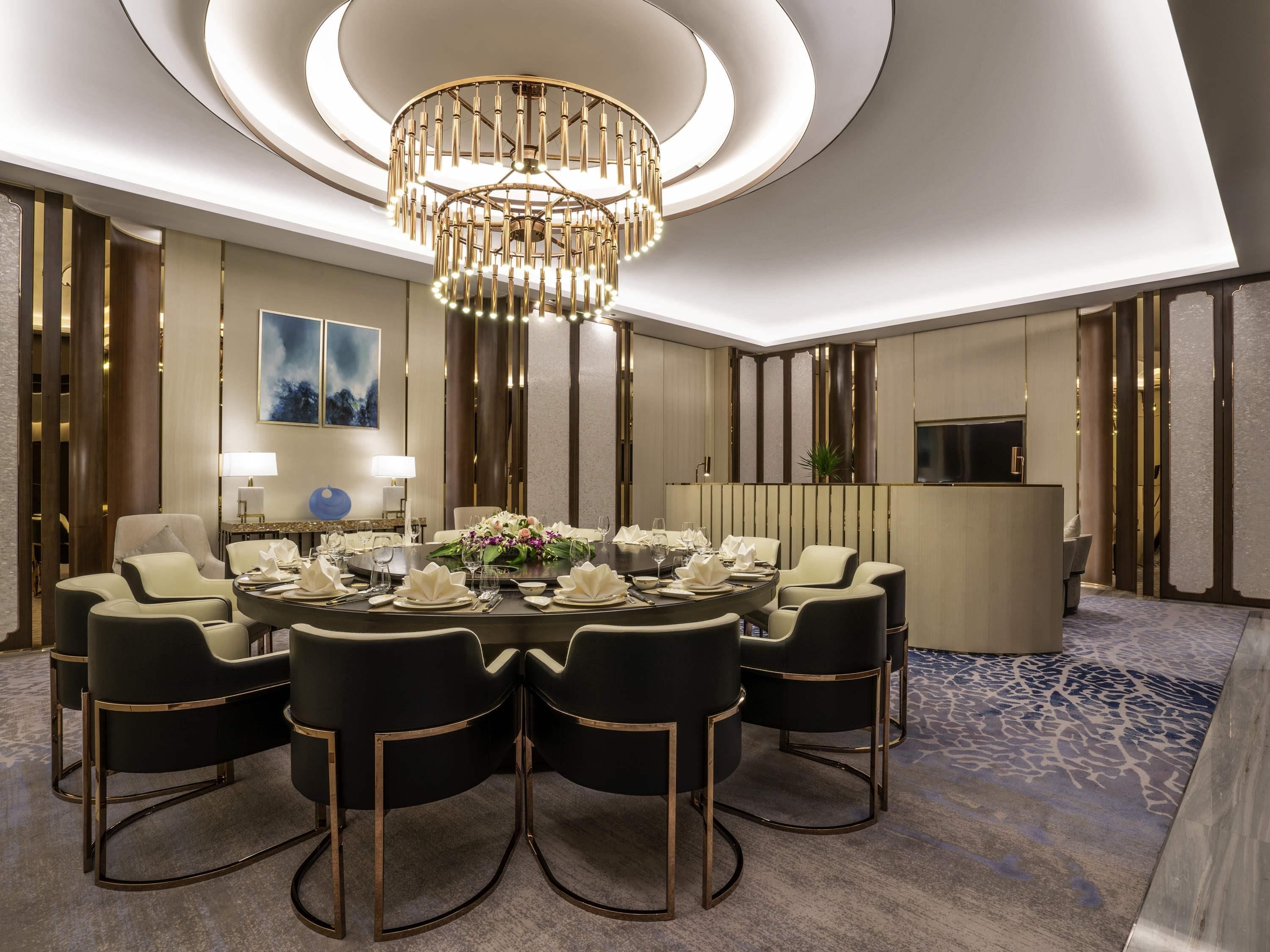 Ein moderner Hotel-Speiseraum mit einem runden Tisch, gedeckt für sechs Personen, umgeben von sechs Stühlen, einer Kristallleuchtre und einem Fernseher an der Wand.