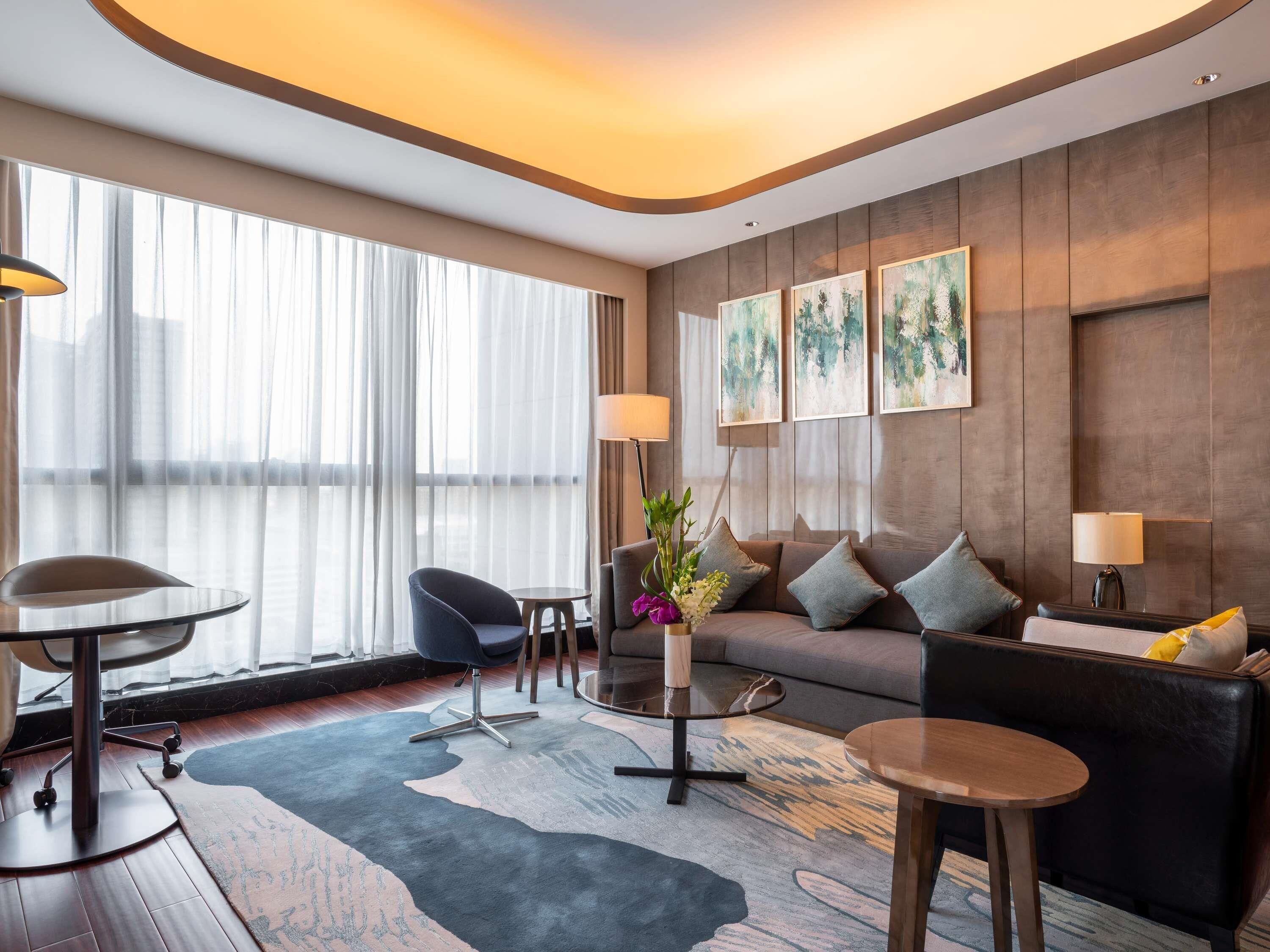 A modern hotel room with a sofa, armchairs, a coffee table, and a dining table. There are large windows with sheer curtains, artwork on the walls, and a wooden floor.